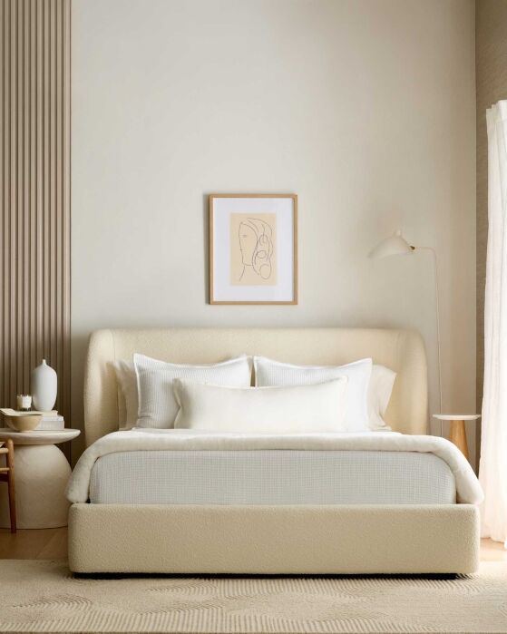 A minimalist bedroom features a cream bed with white linens, a framed abstract line drawing above the headboard, and light-colored decor on side tables.
