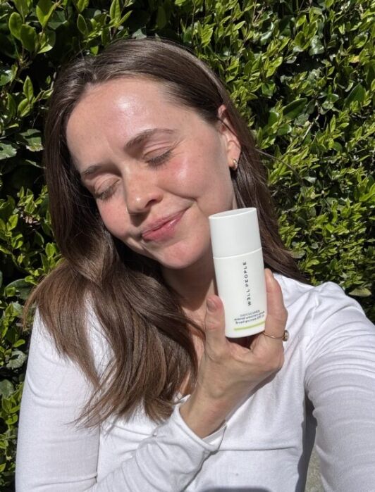 A woman with brown hair holds a white skincare bottle labeled "EVTILORSE" while standing outside in sunlight, smiling with her eyes closed. Green leafy bushes are in the background.