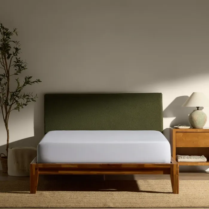 A wooden bed with a green upholstered headboard and white mattress sits against a beige wall, next to a nightstand with a lamp and a potted plant.