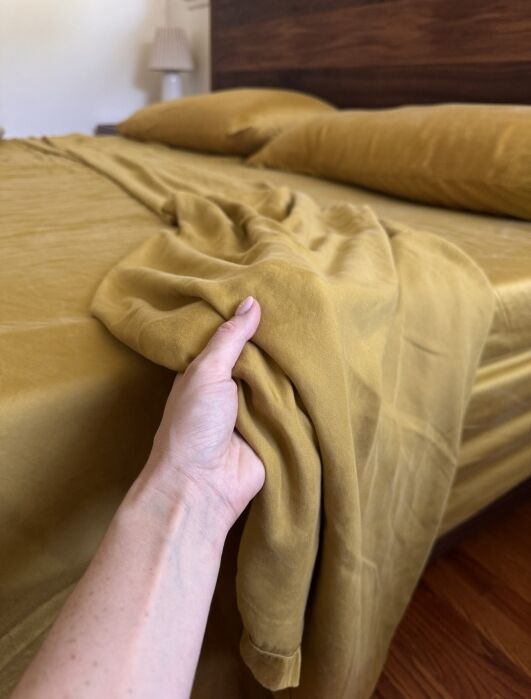 A hand holds the edge of a mustard yellow bedsheet on a neatly made bed with matching pillows and a wooden headboard.