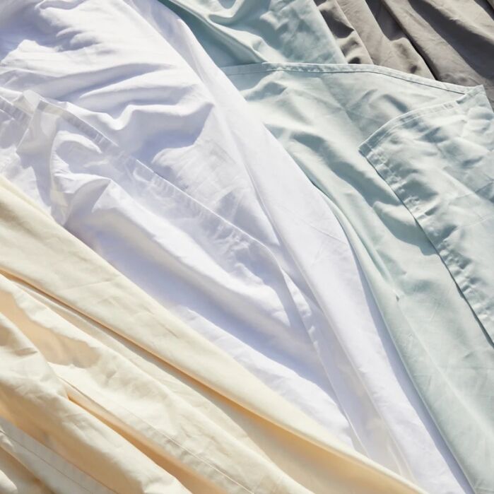 Wrinkled bedsheets in light colors, including white, beige, blue, and gray, are layered and spread out in natural sunlight.