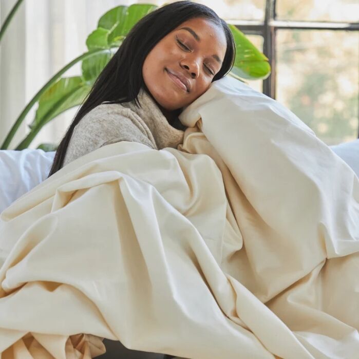 A woman sits on a couch, smiling with her eyes closed, wrapped in a large, cream-colored blanket.