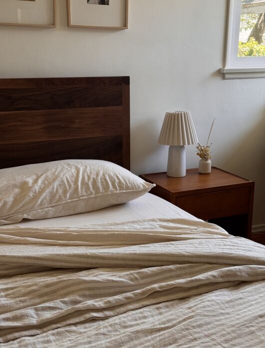 A neatly made bed with beige linens, a wooden headboard, and a nightstand holding a lamp and a small vase, next to a window letting in natural light.