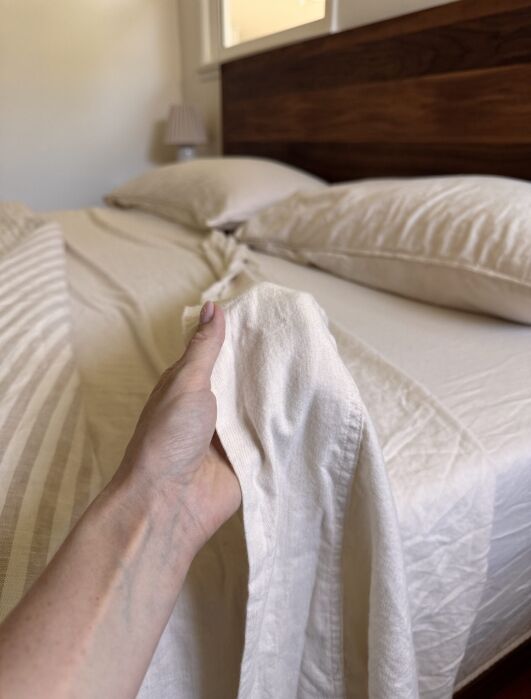 A person’s hand holds a corner of a cream-colored bedsheet while making a neatly arranged bed with matching pillows and a wooden headboard.