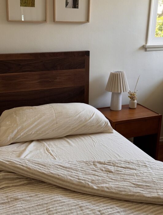 A neatly made bed with a wooden headboard next to a wooden nightstand holding a white lamp and a small vase with dried flowers.