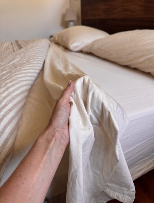 A person’s hand is shown pulling up a bedsheet while making a bed with white pillows and a striped blanket.