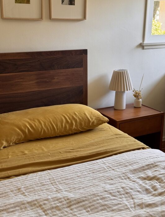 A neatly made bed with a mustard yellow pillow and sheet, a wood headboard, a beige blanket, and a wooden nightstand with a lamp and small dried floral arrangement.