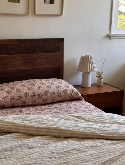 A wooden bed with a floral pillow and beige blanket stands next to a nightstand with a lamp and a small dried flower arrangement under a window.