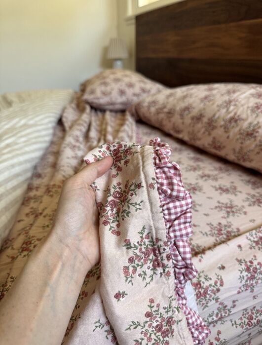 A hand holds the edge of a floral and gingham ruffled quilt on a bed with matching floral bedding and a wooden headboard in a softly lit bedroom.