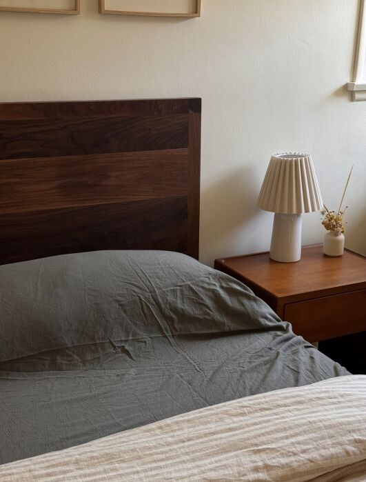 A neatly made bed with a dark wooden headboard beside a wooden nightstand holding a pleated lamp and a small vase with dried flowers.