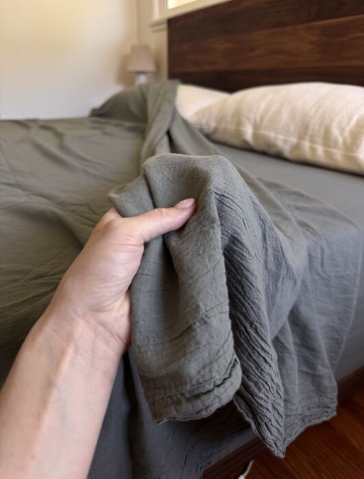 A hand is pulling a gray bedsheet on a neatly made bed with a wooden headboard and a white pillow.