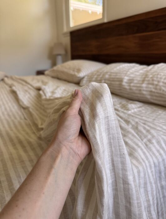 A hand pulling up a beige and white striped bed sheet, with matching pillows and a wooden headboard in the background.