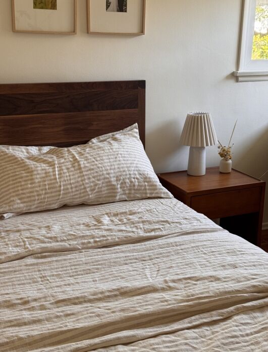 A neatly made bed with striped bedding next to a wooden nightstand holding a lamp and a small vase, under a window and framed artwork on the wall.