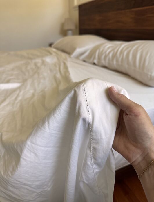 A hand holds the corner of a white bed sheet near a neatly made bed with white pillows and a wooden headboard.