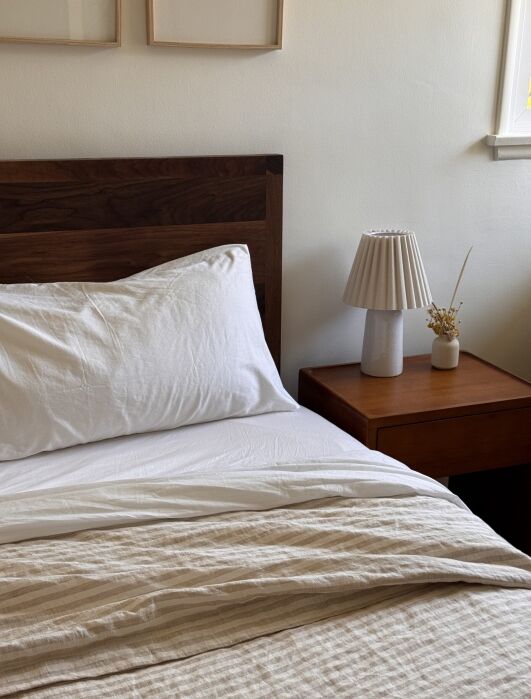 A neatly made bed with white linens next to a wooden nightstand holding a small lamp and a vase with dried flowers in a softly lit room.