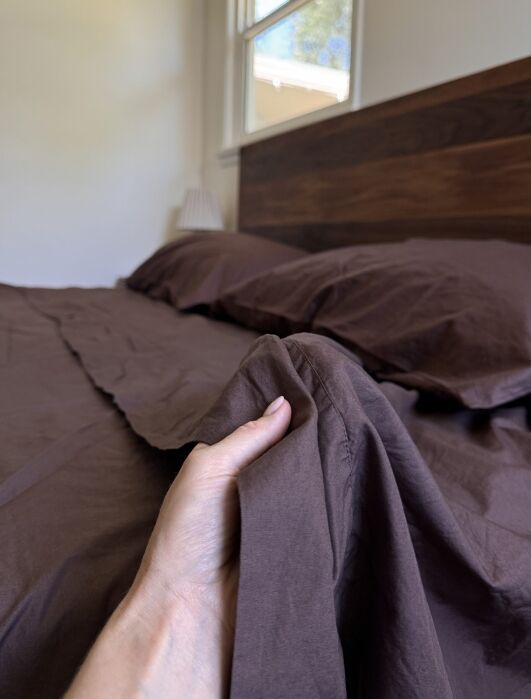 A hand holding the edge of a brown sheet on a neatly made bed with matching brown pillows, next to a wooden headboard and a window.