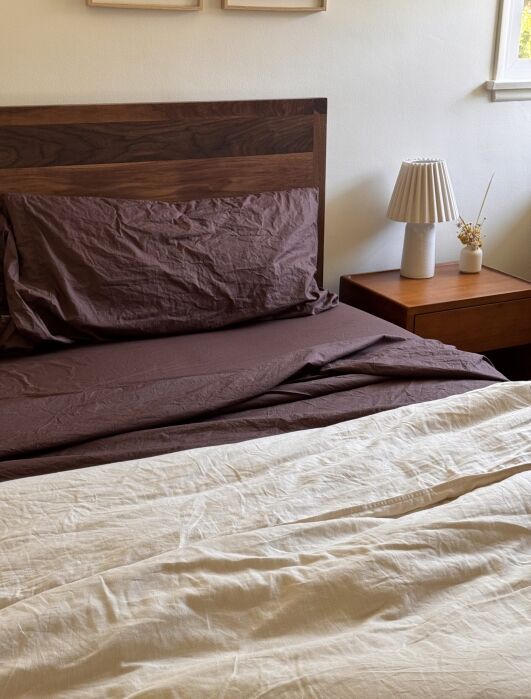 A neatly made bed with brown sheets and pillow, a wooden headboard, a nightstand with a lamp, and a small vase, next to a window with natural light.