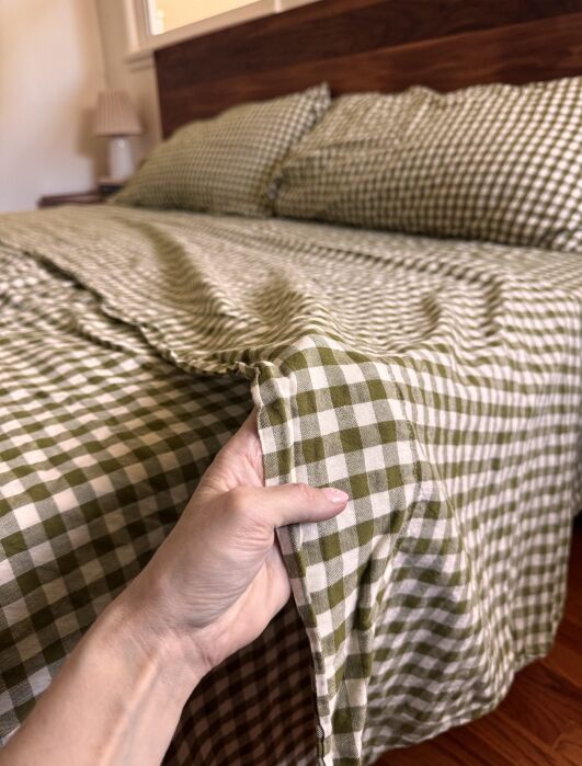 A hand holds the corner of a green and white checkered bedspread on a neatly made bed with matching pillows and a wooden headboard.