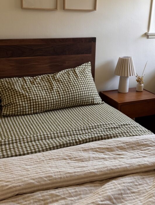 A neatly made bed with green checkered bedding, a wooden headboard, a matching wooden nightstand, a small lamp, and a dried floral arrangement.