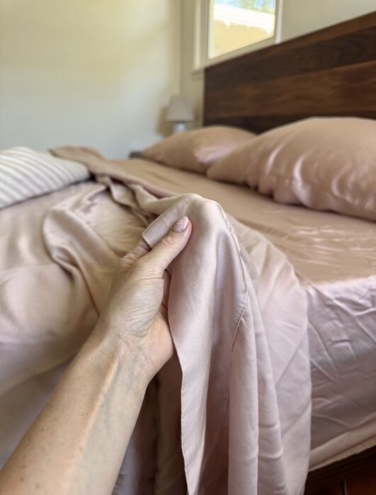 A hand holds the edge of a light pink silky bed sheet, with a made bed, pillows, and headboard visible in the background.