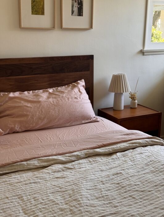 A wooden bed with pink bedding sits next to a wooden nightstand holding a lamp and small vase; two framed pictures hang on the wall above.