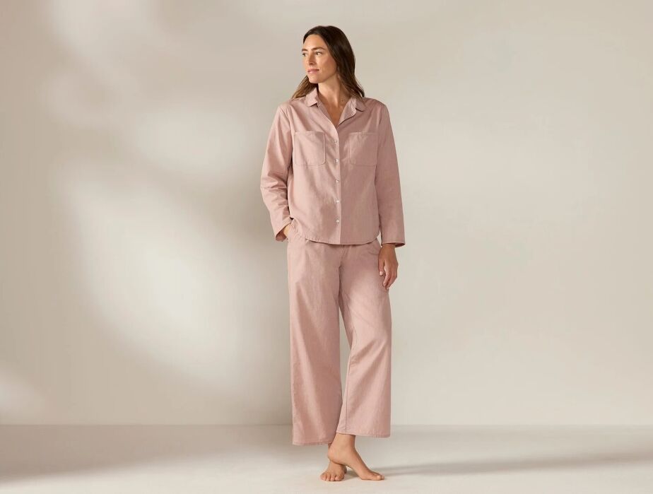 A woman stands barefoot against a plain background, wearing light pink long-sleeved pajama top and pants.