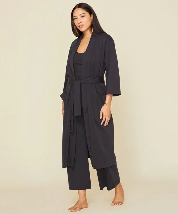 A woman stands barefoot wearing a black belted robe over matching black pants and a top, posed against a plain beige background.