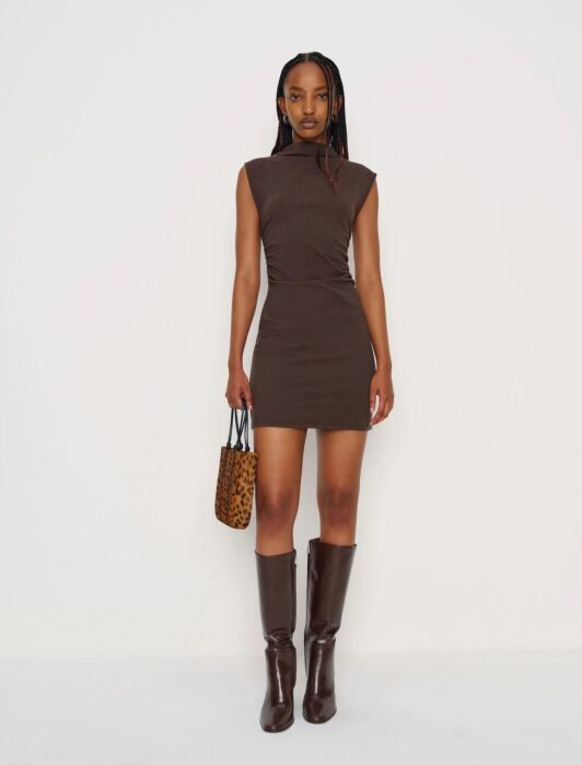 A woman in a sleeveless brown mini dress stands against a white background, holding a leopard print handbag and wearing knee-high brown boots.