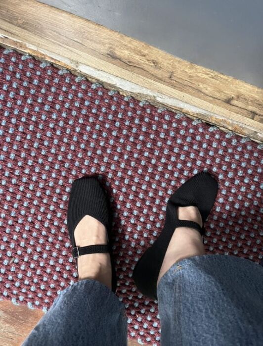 A person wearing black Mary Jane shoes and blue jeans stands on a red and blue patterned carpet near a wooden threshold.