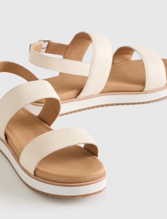 A pair of beige sandals with double straps and ankle buckle, featuring a cushioned tan footbed and white soles on a light background.