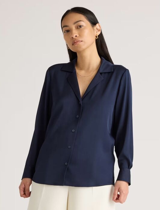 A woman stands facing forward wearing a navy blue button-up blouse with a collar and long sleeves, paired with off-white pants against a plain background.