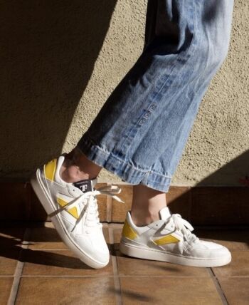 A person wearing blue jeans and sustainable sneakers with white and yellow accents stands on a tiled floor in sunlight.