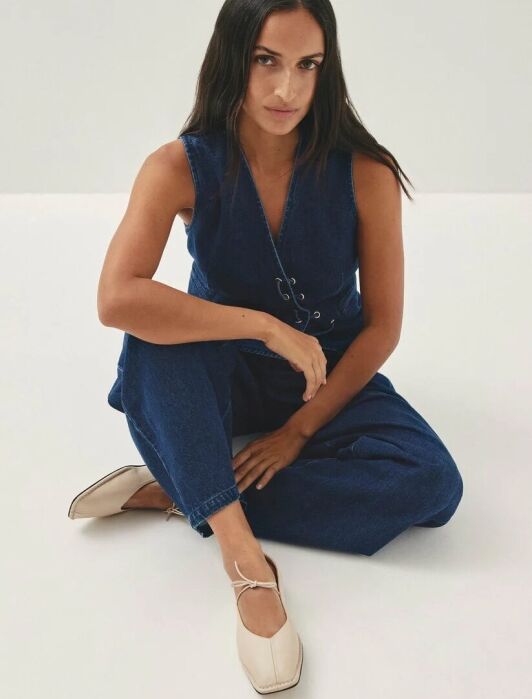 A woman with long dark hair sits on the floor wearing a sleeveless denim jumpsuit and beige square-toe flats, looking at the camera.