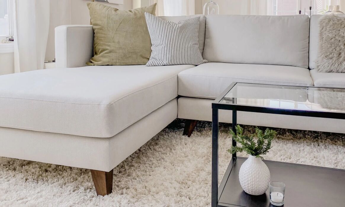 Modern living room with a white sectional sofa, green and striped pillows, glass coffee table, small plant, candle, and a textured white rug.