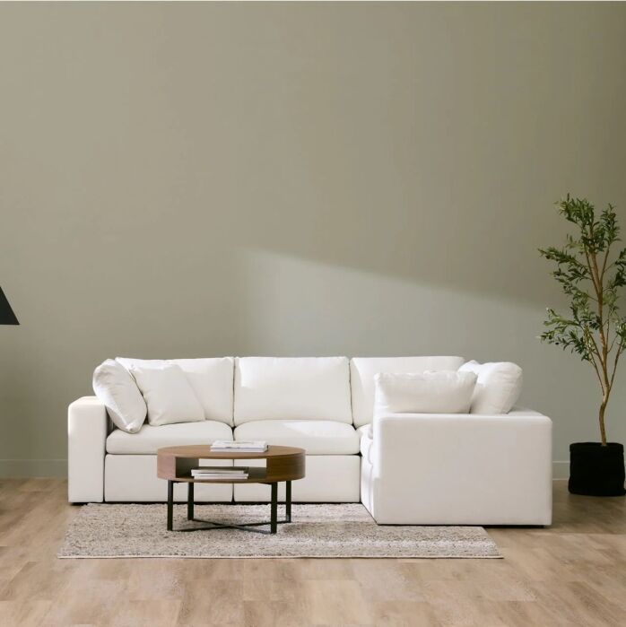A white sectional sofa sits on a light rug with a round wooden coffee table in front, next to a tall plant, against a pale wall in a minimalist room with wood flooring.