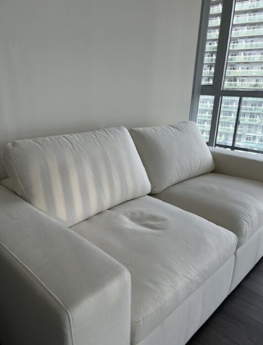 A white two-seater couch sits against a plain wall near a window with blinds, letting in daylight.