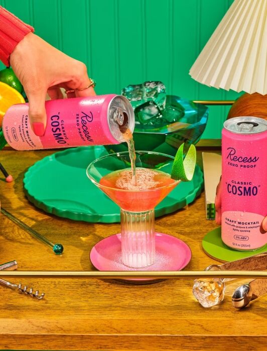 A can of Recess Classic Cosmo is poured into a cocktail glass on a wooden table with limes, a green tray, an ice bucket, lamp, and bar tools.