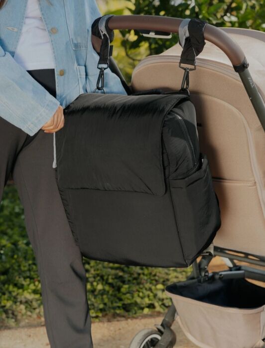 A person stands beside a beige stroller with a large black bag attached to the handlebar using two straps.