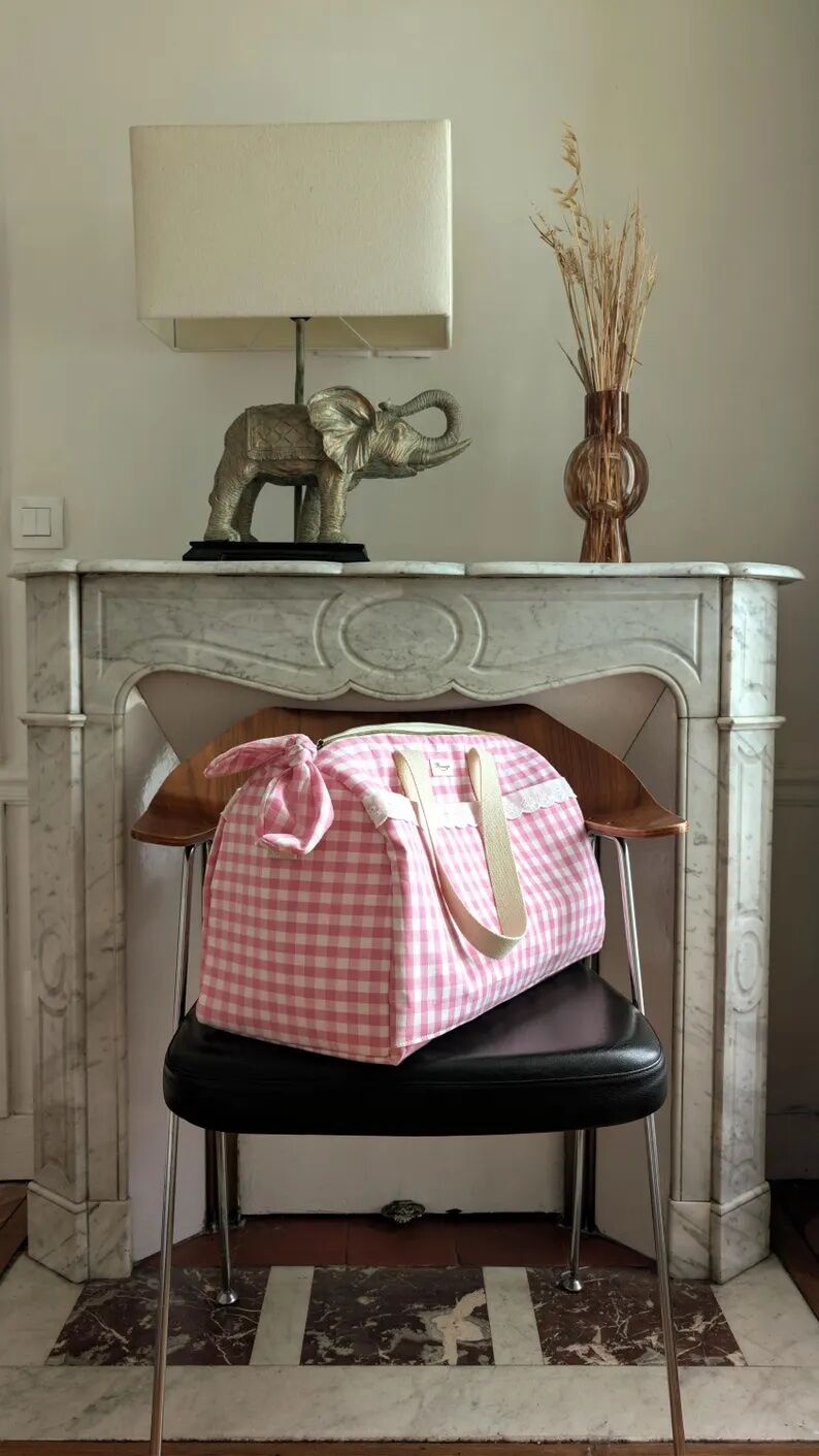 A pink gingham duffel bag sits on a black chair in front of a marble fireplace with a lamp, an elephant figurine, and a vase of dried grasses.