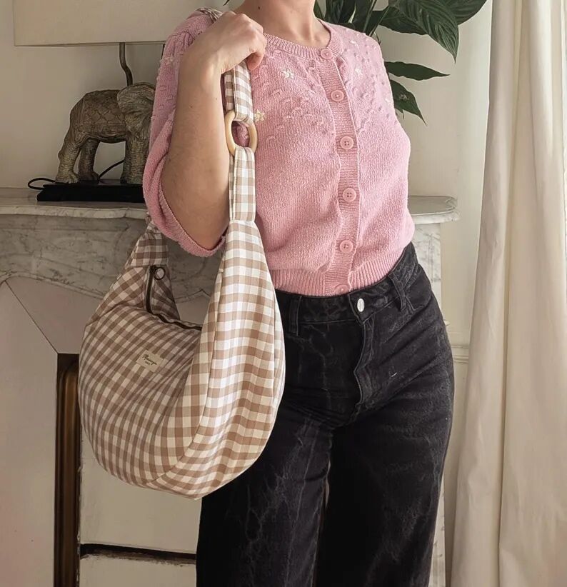 Person wearing a pink knit cardigan and black jeans holds a large beige and white checkered shoulder bag while standing indoors near a marble surface with a decorative elephant.
