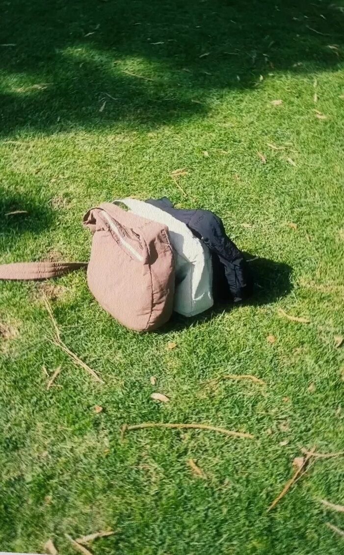 A tan messenger bag and two pieces of clothing, one light and one dark, lie on green grass in sunlight.