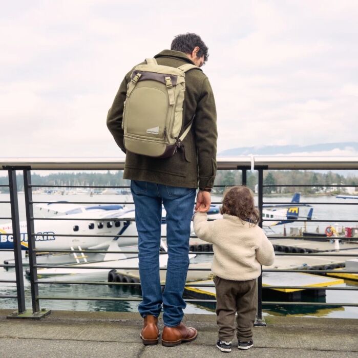 An adult and a child stand on a waterfront promenade, holding hands and facing floatplanes docked on the water.