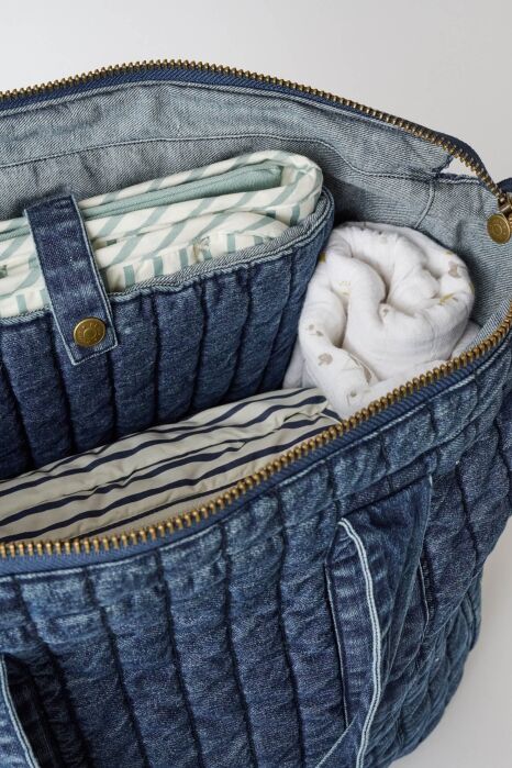 Open denim bag containing a folded striped mat, a rolled white blanket with small prints, and a striped fabric item, all neatly arranged inside.