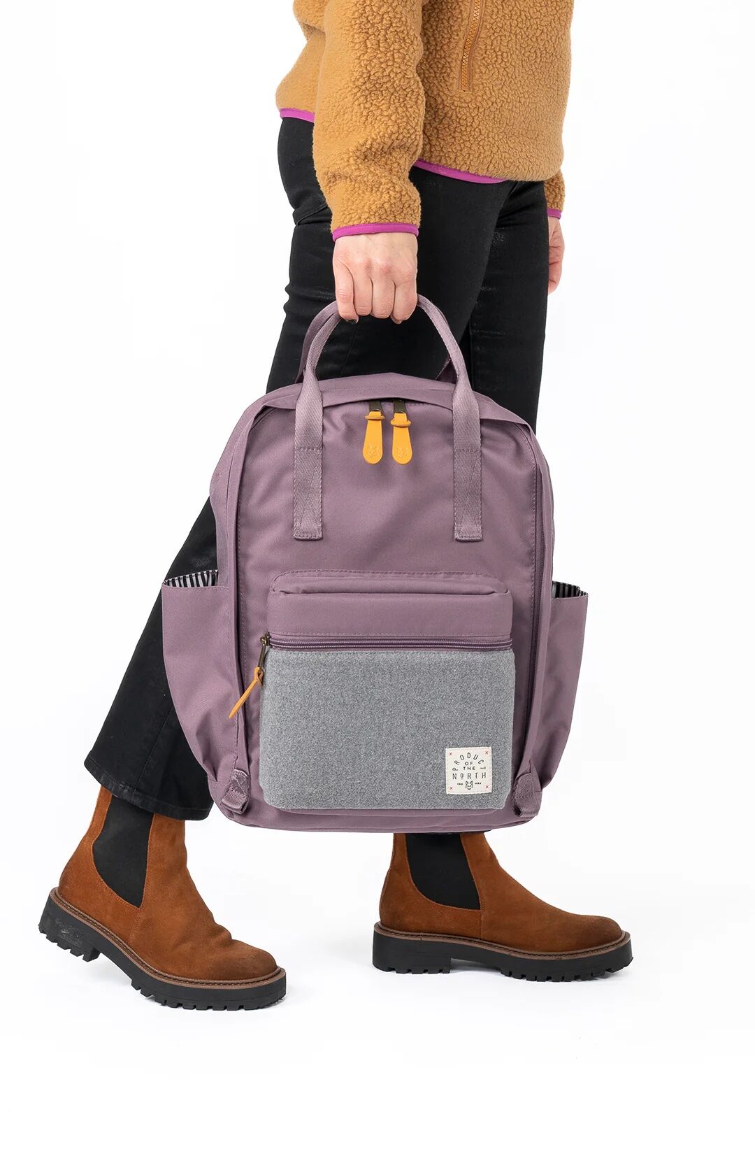 Person wearing brown boots and black pants holds a lavender backpack with grey front pocket and orange zipper pulls against a white background.