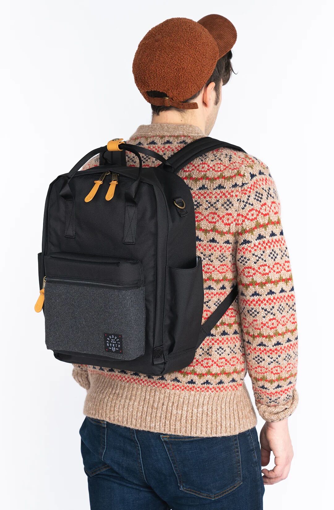 A person wearing a brown cap, patterned sweater, and jeans is seen from behind, carrying a black and gray backpack with tan zipper pulls.