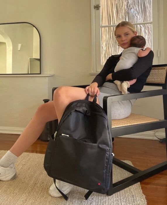 A woman sits on a chair holding an infant on her shoulder, with a black backpack by her side in a softly lit room.