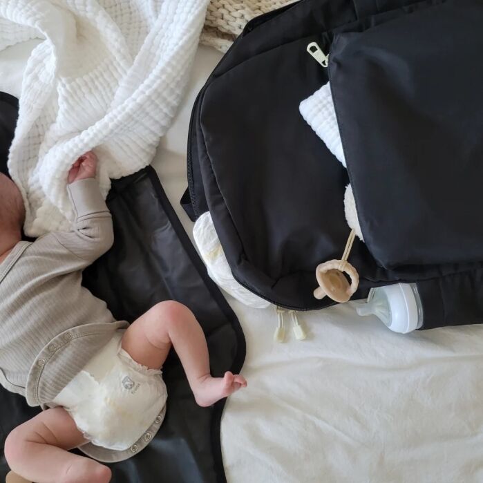 A baby in a diaper lies on a changing mat next to a black diaper bag containing a baby bottle, pacifier, and white blanket.