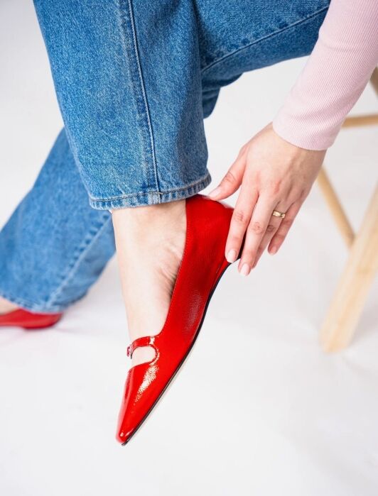 Person wearing blue jeans and a light pink top slips on a shiny red flat shoe while seated on a wooden stool.