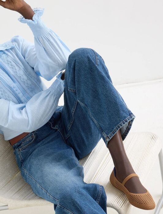 A woman with braided hair sits on a chair, wearing a light blue blouse, blue jeans, and brown flats, against a plain white background.