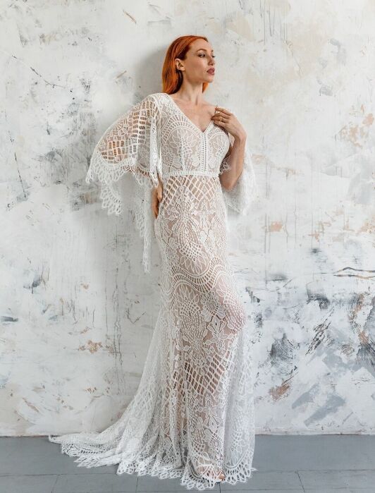 A woman with red hair poses in a long, white, lace dress with intricate patterns, standing against a textured, abstract white and gray wall.